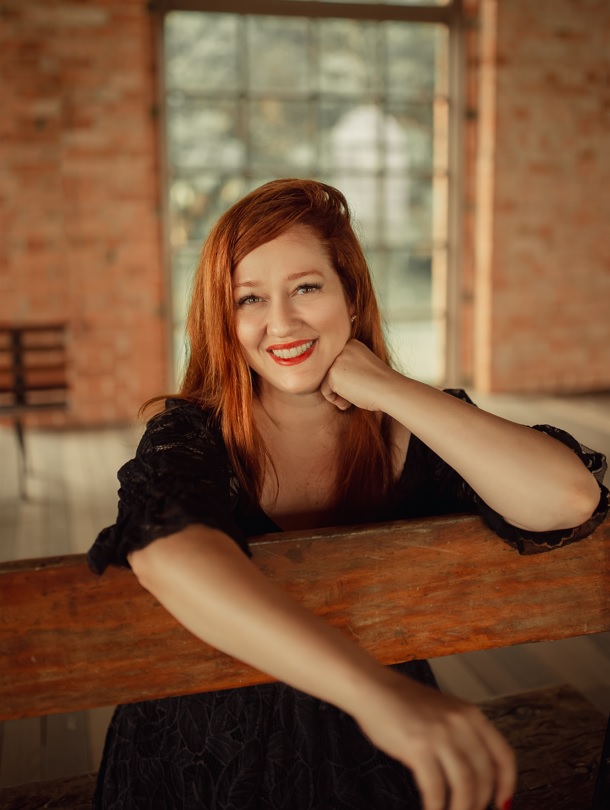 Rachel das Flores sorrindo, apoiada em um banco de madeira em ambiente rústico com parede de tijolos